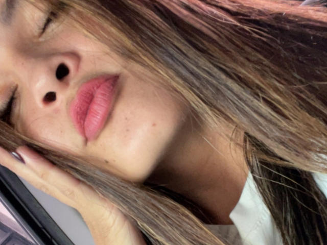 Gabrielafoxxy's face, brown hair, pink lips, nose, and hand touching her head, showing a slight smile with eyes closed. She's wearing a white shirt.