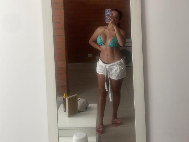 Gabrielafoxxy in a green bikini top and white shorts, standing in front of a mirror, holding a phone, with a tan, toned body and small pink slippers.