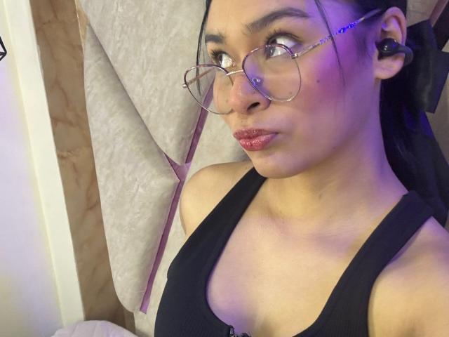 IsaDoux with round glasses, black tank top, and shiny pink lipstick, looking to the side with a slight pout, earbuds in, purple light on her face.