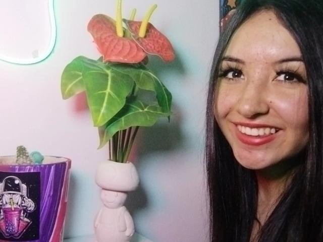 SusanMonroy with long black hair, smiling, has freckles, and is wearing red lipstick. She's next to a white vase with green leaves and pink flowers, and a purple cup.