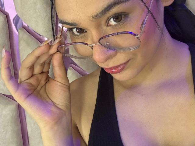 IsaDoux with glasses, holding them with her right hand, has brown eyes, pink glossy lips, and black tank top, showing her boobs slightly, with dark hair.