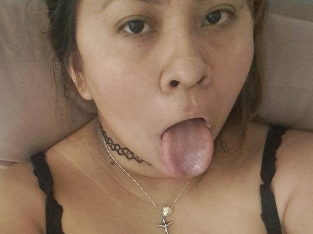 ValeriaFranco with her tongue out, staring at the camera, wearing a black lace top, choker necklace, and cross necklace, with her brown hair down and slightly messy.