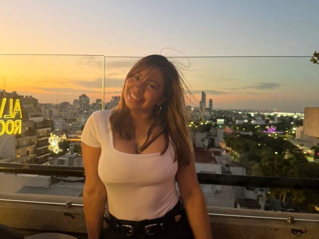 ValeriaFranco with medium tits, wearing a white, tight, short-sleeve top and black pants, smiling on a rooftop at sunset, city lights in the background, glass railing in front.