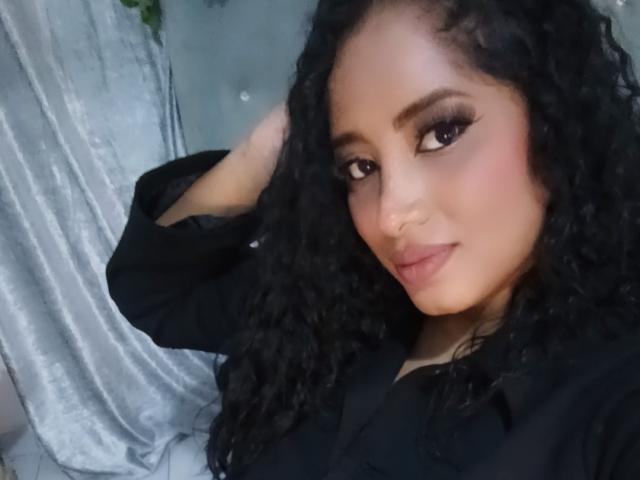 MariaCatalina with curly black hair, wearing a black shirt, has a nose ring and is smiling with dark, dramatic eyeliner. Her hand is in her hair.