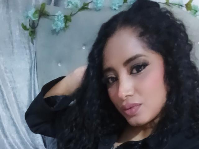 MariaCatalina with curly black hair, dark eyeliner, and pink lipstick, wearing a black shirt, looking at the camera with a slight pout, hand in her hair.