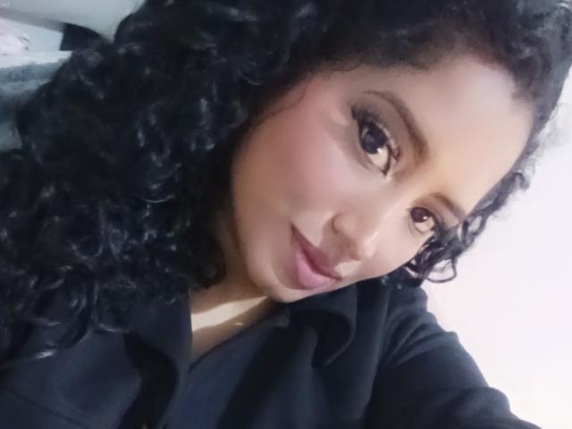 MariaCatalina has curly black hair, brown eyes, and is wearing a black shirt. She's smiling with pink lipstick and a slight nose ring. Her ass is not visible.