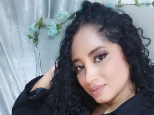 MariaCatalina with curly black hair, dark eyes, and full pink lips, wearing black, looking slightly to the right, soft smile, subtle makeup, gray background with blue flowers.