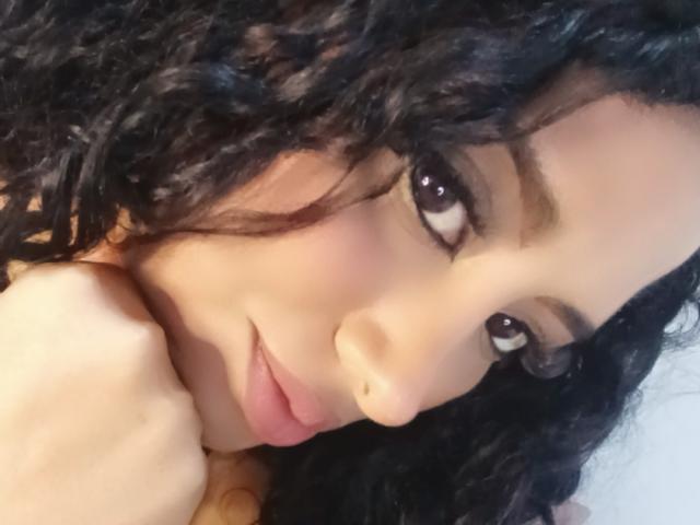 MariaCatalina with curly black hair, brown eyes, and pink lips, looking at the camera with a slight smile, her hand near her face, soft light on her skin.