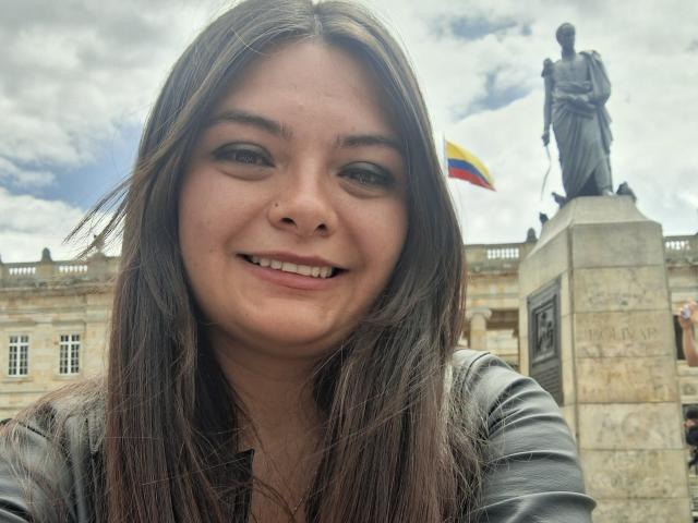 katherinLatin with long brown hair, smiling, nose ring, wearing a gray leather jacket, standing in front of a statue, Colombian flag in background, cloudy sky.