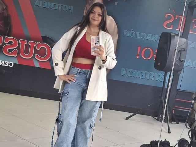 katherinLatin with long brown hair, red crop top, white coat, and blue cargo pants, taking a selfie in a room with a microphone stand and speaker.