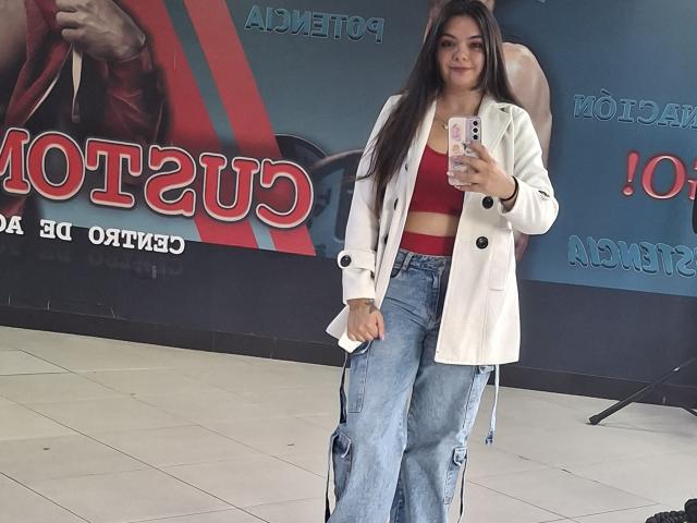 KatherinLatin with long dark hair, wearing a white jacket, red crop top, and high-waisted blue jeans, holding a phone, standing in a tiled room.