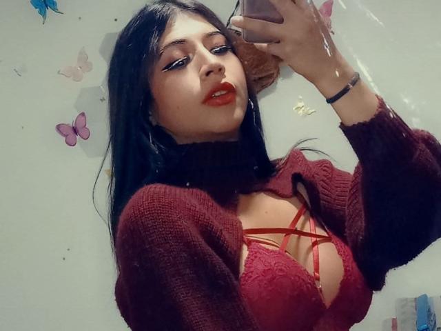 MissCristina69 with red lipstick, black hair, wearing a maroon knit top over a red lace bra with strappy detail, taking a mirror selfie with her phone.