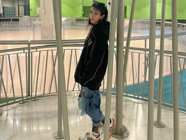 LaurenEvie69 with long dark hair, black hoodie, blue pants, and white sneakers with red stripes, standing under metal poles in a modern, bright indoor space.