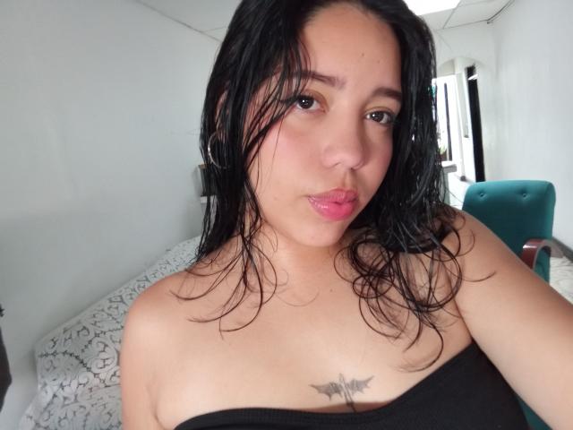 Lila, con el pelo negro y húmedo, labios rosados y un top negro sin tirantes, mostrando un pequeño tatuaje de murciélago en el pecho, mirando a la cámara..