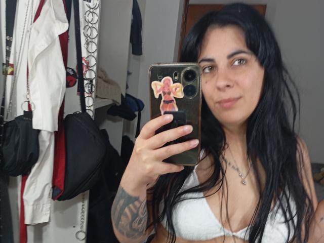 SindyLove with black hair, white bra, tattoo on right arm, holding phone with Minnie Mouse sticker, mirror selfie, slight smile, gray eyes, white walls, black bag, white robe.