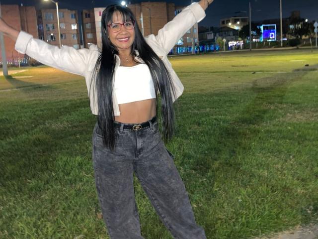MarcelaHotty with long black hair, glasses, white crop top, beige jacket, and dark jeans, arms spread, smiling, standing on grass at night, her small tits visible.