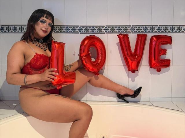 DanniLuke in red lace lingerie, holding red love balloons, sitting in a white bathtub, wearing black heels, with tattoos on her thigh and wrist, dark makeup, and black eyeliner.