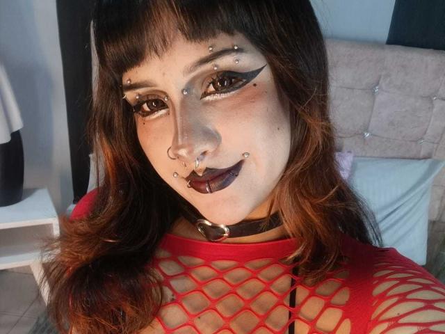 DanniLuke with dark makeup, nose piercings, and facial piercings, wearing a red fishnet top, black choker, and dark lipstick, has brown hair and a cute face.