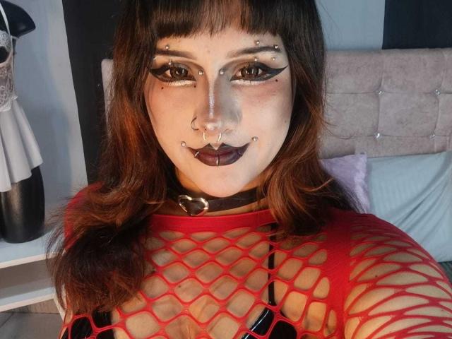 DanniLuke with dark makeup, septum ring, and black lipstick, wearing a red fishnet top, revealing her tits, and a black choker with a heart buckle.