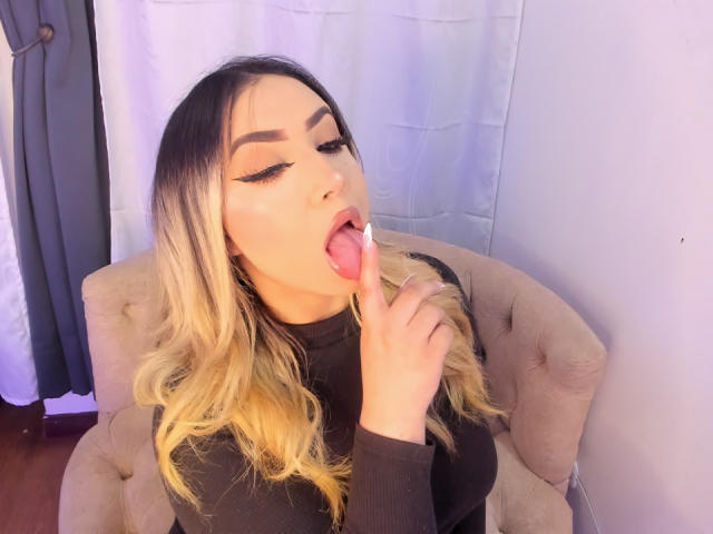 Melissa Lust, avec ses cheveux blonds dégradés, son eye-liner noir et ses sourcils bien définis, la langue tirée et léchant son doigt, portant un haut noir, assise sur une chaise beige capitonnée..