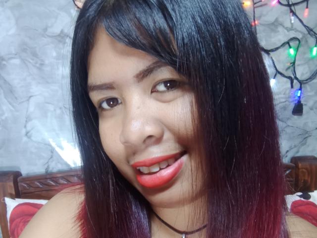 MariahSquirting69 with black and red gradient hair, red lipstick, smiling, brown eyes, and light skin, showing teeth, in a selfie with Christmas lights in the background.