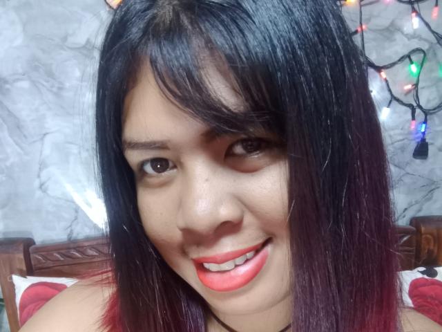 MariahSquirting69 has black hair with purple tips, brown eyes, and a red lipstick smile. She's wearing a red top, with colorful Christmas lights in the background.