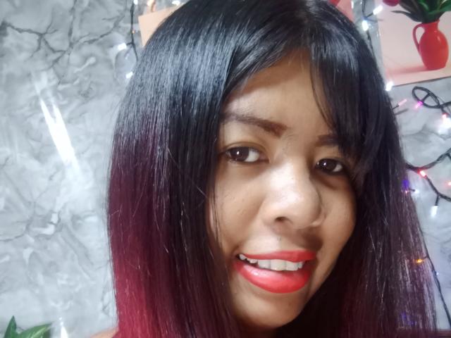 MariahSquirting69 with dark, glossy black-to-red ombre hair, smiling with red lipstick, brown eyes, and medium brown skin, showing teeth. Background has string lights and a red vase.