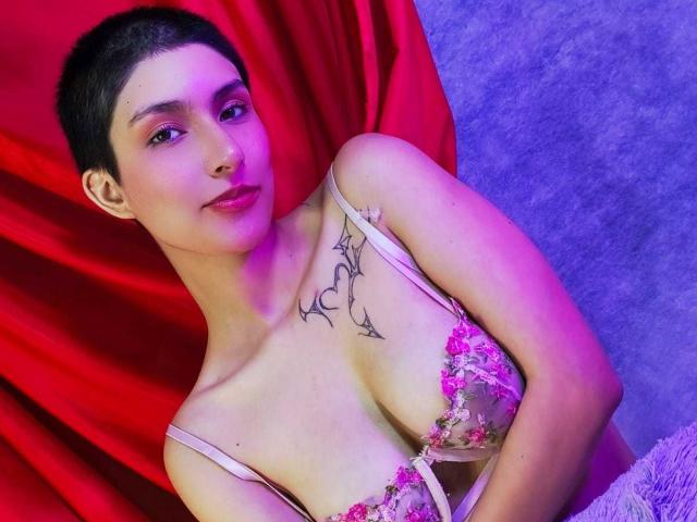IsabellaHallad with short black hair, pink floral bra, small tattoo above tits, purple-pink lighting, red curtain, grey carpet, slight smile, confident pose.
