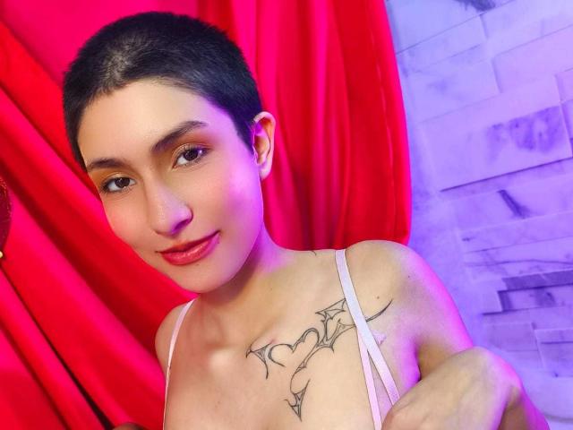 IsabellaHallad with short black hair, red lipstick, and a tattoo on her chest, wearing a pink top, smiling at the camera. Red curtains and wooden floor in background.