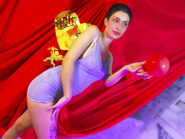 IsabellaHallad in a tight, light purple dress, holding a red balloon, has short dark hair, a tattoo on her thigh, and a golden mask on her back. Red curtain.