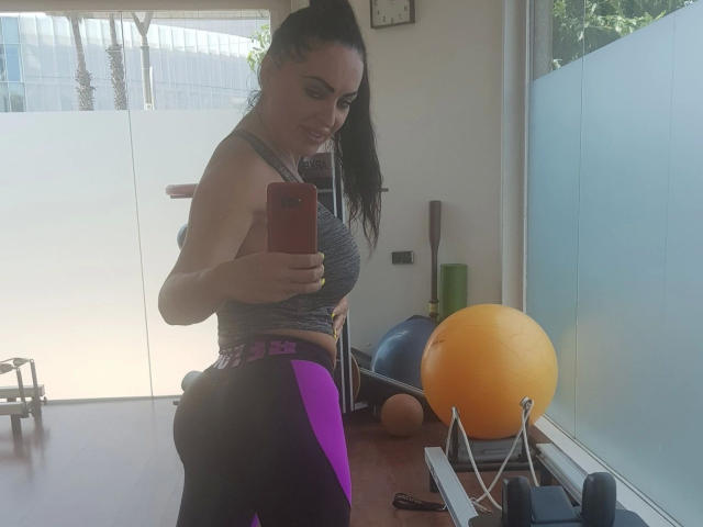AnnaLust1986-hot, standing in a gym, takes a mirror selfie. She has a toned, athletic build with a prominent, rounded butt in purple and black workout pants. She wears a gray sports bra and has long, dark hair in a high ponytail. She holds a red smartphone in her right hand. In the background, there's a yellow exercise ball, a blue stability ball, and a wooden pole. Bright, natural light from a window on the right.