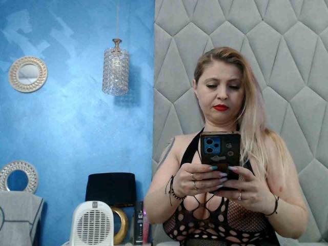 AretiAlisya with blonde hair, red lipstick, and black mesh top showing her large tits, holding a phone, standing in a room with blue walls and a quilted grey wall.