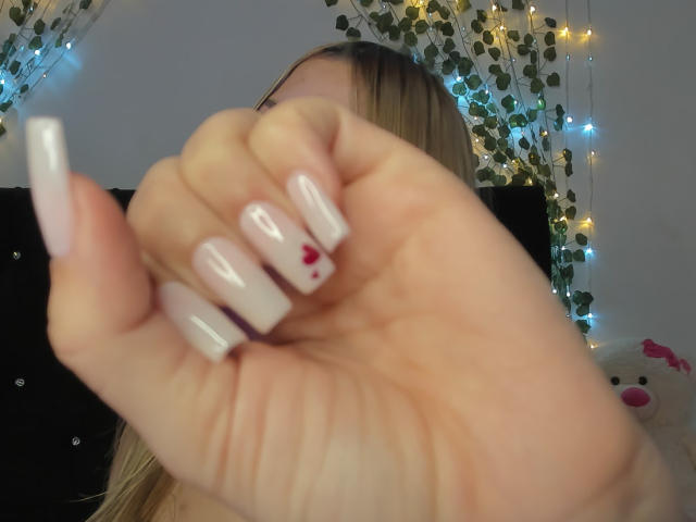 La main de ShantalSummer, avec des ongles blancs et brillants et des points rouges sur l'ongle de l'index, tenant une lime à ongles blanche, dans une photo floue prise en contre-plongée..