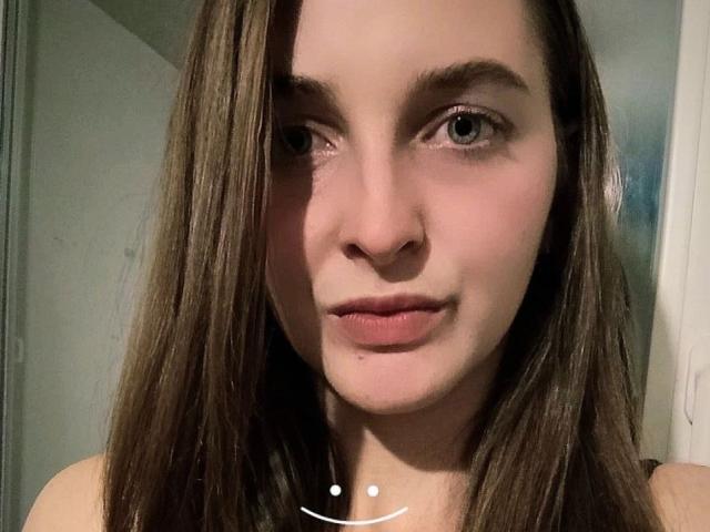 Close-up portrait of SlavyanaReshh with light skin and brown, straight hair falling to the sides. She has blue eyes, thin eyebrows, and pink lips. A small pimple is visible on her lower chin. Her expression is neutral. She wears a thin, white necklace with a small, circular pendant. The lighting is soft, highlighting her facial features. The image is slightly shadowed on the left side.