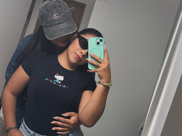 A selfie of KymAndNala in a bathroom. KymAndNala, with tan skin and dark hair, wears a black t-shirt with a Hello Kitty logo and blue jeans. She holds a green phone in her right hand. A person with tan skin, wearing a gray denim cap, stands behind her, hands on her hips. KymAndNala has a black eye mask and a gold bracelet. The cap has a small white outline of a heart. The image is casual and modern.