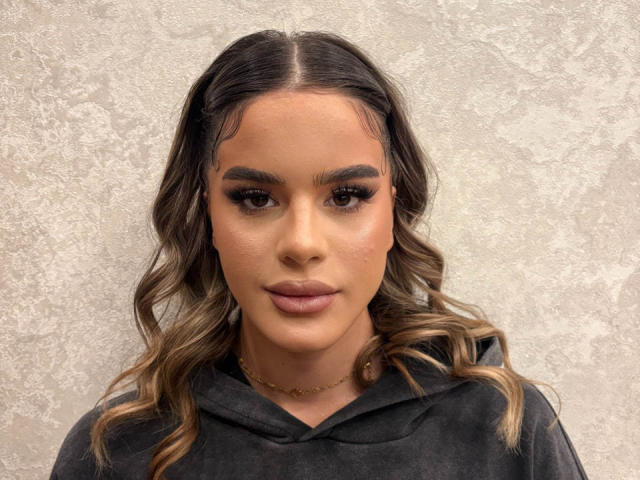 NikkiPearl has wavy, shoulder-length brown hair with blonde highlights, parted in the middle. She has dark eyebrows, long eyelashes, and full, pink lips. She wears a dark grey hooded shirt and a gold chain necklace. Her skin is tan and smooth. The texture of the wall behind her is rough and patterned. The image focuses on her face, which is centered and slightly tilted to the left. Her expression is neutral. The overall style is modern and straightforward.