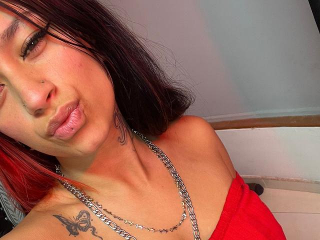 Close-up of RoxanaSanchezz with medium-length, dark red-brown hair. She has a small nose ring on her right nostril and a butterfly tattoo on her left shoulder. RoxanaSanchezz is wearing a red strapless top and a silver chain necklace. Her lips are puckered in a subtle kiss. Her skin is light brown, and she has minimal makeup. The lighting is bright, highlighting her facial features and the texture of her hair. The image focuses on her upper torso and face.