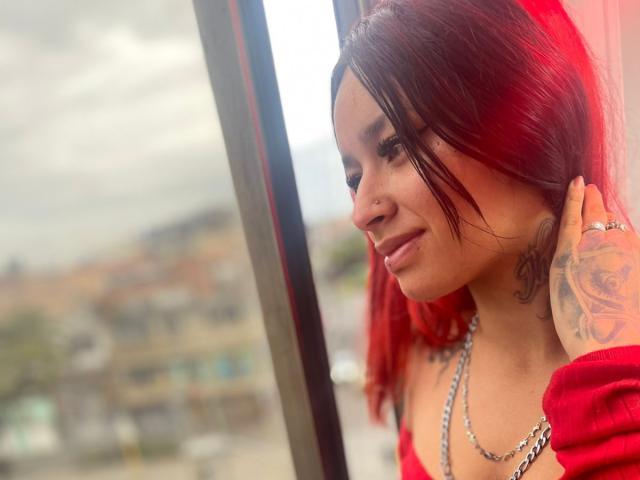 RoxanaSanchezz, a young woman with vibrant red hair, stands by a window. She has a small nose ring in her left nostril and a tattoo on her right hand. She wears a red off-shoulder top, a silver chain necklace, and has dark eyeliner. Her right hand is touching her hair. The blurred background shows a cloudy sky and urban buildings. Her skin tone is light, and she has a confident, slightly relaxed expression. The photograph emphasizes her facial features and hair color.