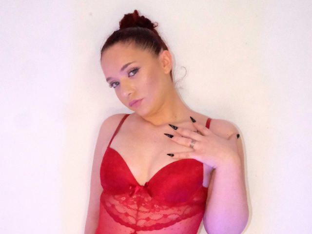 BaddHabits with red lace bra, showing her tits, black nails, hand on chest, hair in bun, white wall, soft pink makeup, and ring on her finger.