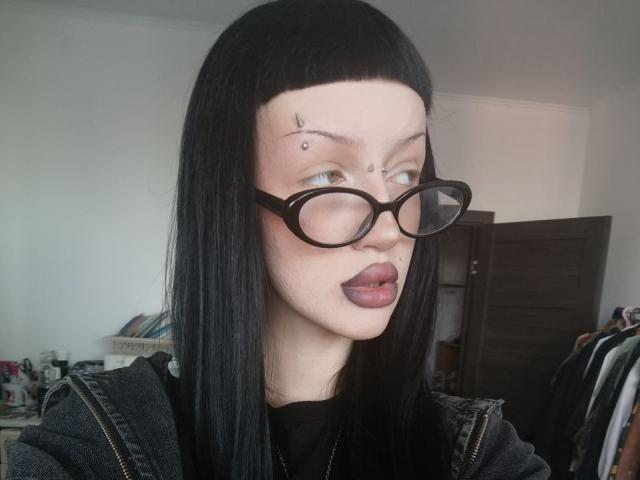 A close-up photo of SexsyNanita with long black straight hair and blunt bangs. She wears black-rimmed glasses, has dark purple lipstick, and a small star tattoo on her right forehead. Her mouth is slightly open. She's dressed in a black shirt with a denim jacket. The background is a slightly blurred room with a dark wooden door and a shelf with clothes. The image has a realistic, candid style.