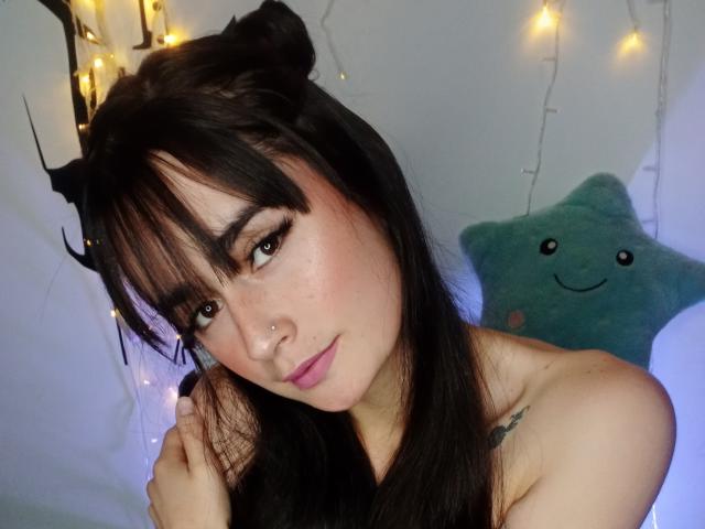 EllieBonneth with dark hair in double buns, brown eyes, pink lips, nose ring, and freckles, looking at camera, topless, small tattoo on right shoulder, soft smile.