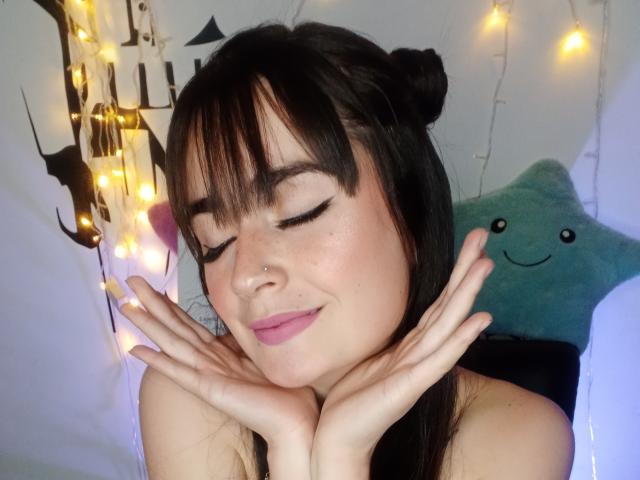 EllieBonneth with her eyes closed, pink lipstick, black hair in buns, nose piercing, and hands near her face, smiling softly. Fairy lights and a green star pillow behind her.