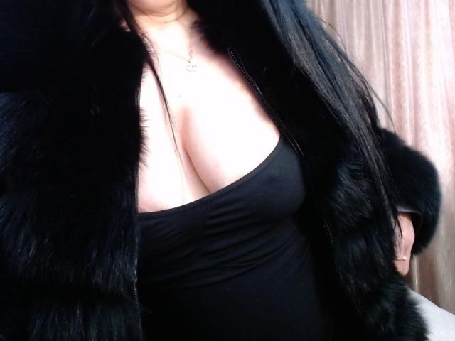 SiPassioneX with large boobs, wearing a black fur vest over a black tank top, her long black hair partially visible, a small necklace with a heart charm.