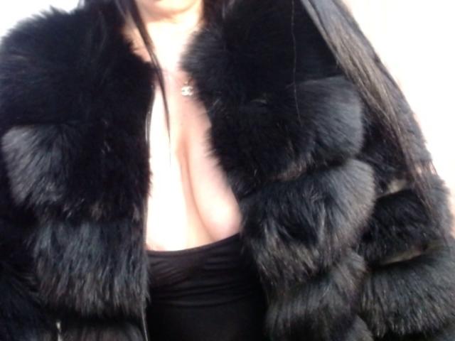 SiPassioneX is wearing a black, fluffy fur coat with a deep cleavage, exposing her tits. She has long, straight black hair and is wearing a black top.