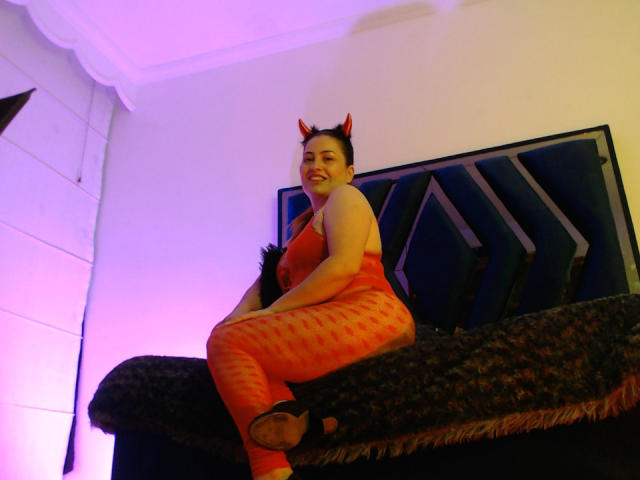 DayanHank vestida con un traje rojo estampado de una sola pieza, con cuernos de diablo, sentada en un sofá oscuro y peludo, sonriendo, con orejas de gato negras y luces moradas en el fondo..