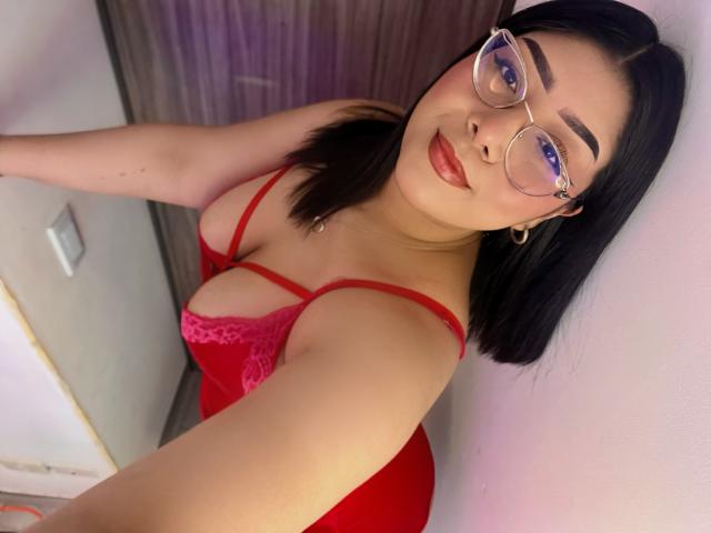 AlexaKiim, a young Asian woman with straight black hair, wears glasses with purple-tinted lenses. She is dressed in a red lace bralette that highlights her cleavage. Her skin is smooth and light-toned. She has full lips with red lipstick and is looking slightly upward at the camera. She has a small silver necklace and gold hoop earrings. The image is a close-up selfie, showing her upper body and face. The background is blurred.