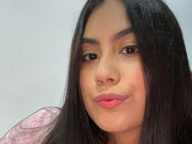 CurvyBuff has long black hair, brown eyes, and pink lipstick. She's smiling with slightly flushed cheeks, wearing a pinkish top. Her nose has a small beauty mark.