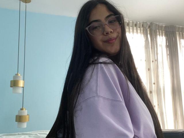 CurvyBuff with long black hair, glasses, and pink lipstick, wearing a purple shirt, smiling at the camera, showing off her cute ass in a cozy room.