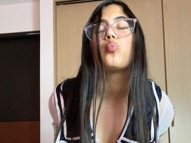 CurvyBuff with long black hair, wearing glasses and a low-cut top, puckering her lips in a kissy face. Her tits are partially visible, with a slightly open top.