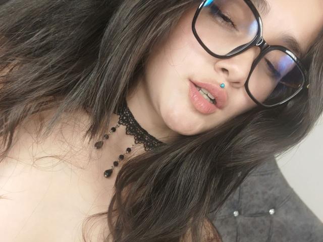 Close-up of NataliaRivas with long, wavy dark brown hair. She wears black lace choker with black bead accents and black-framed glasses. Her lips are slightly parted with a blue lip piercing. Her skin is fair, and she has a relaxed, slightly seductive expression. The background is blurred and dark. The image has a soft, natural light. Her glasses have a slight reflection on the lenses. The choker's lace texture contrasts with her smooth skin. The overall composition is intimate and detailed.